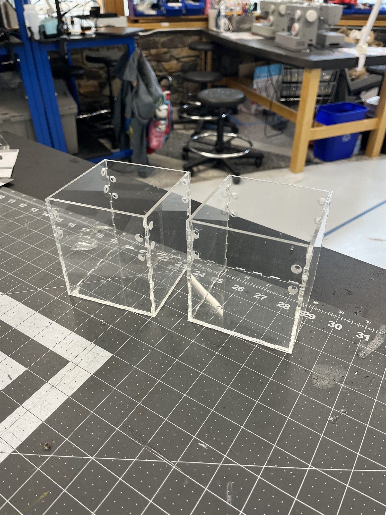 Two laser-cut acrylic box housings standing on a cutting mat in the shop, bolt holes along their edges.