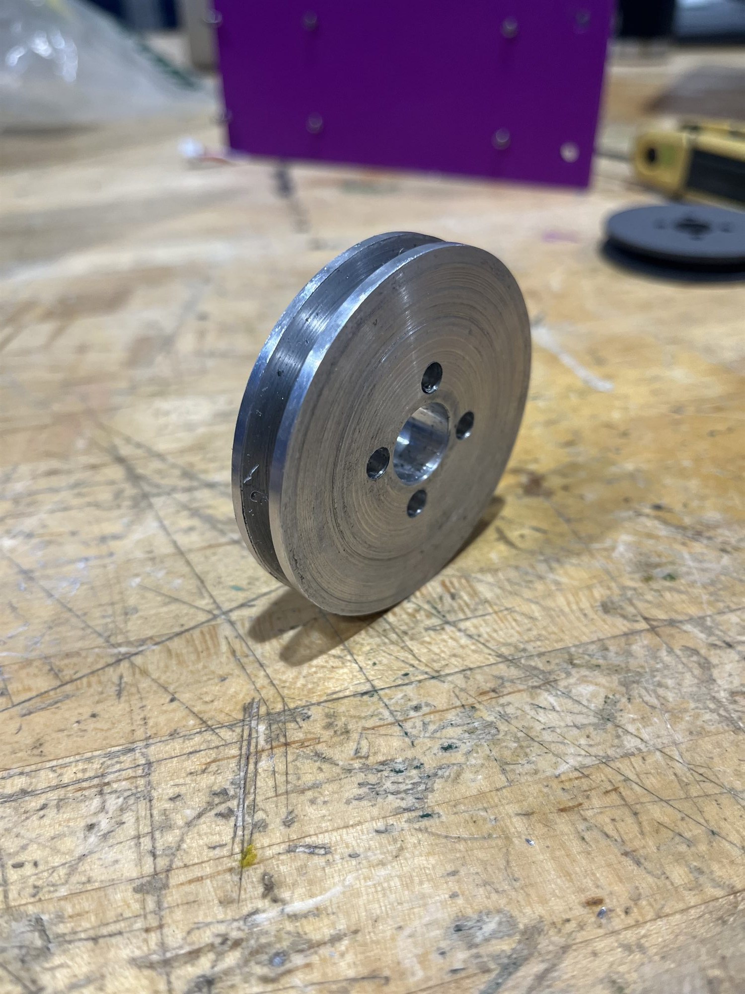 A pulley turned from 6061 aluminum standing on a workbench, lathe tool marks visible across the face, with its V-groove and bolt circle.