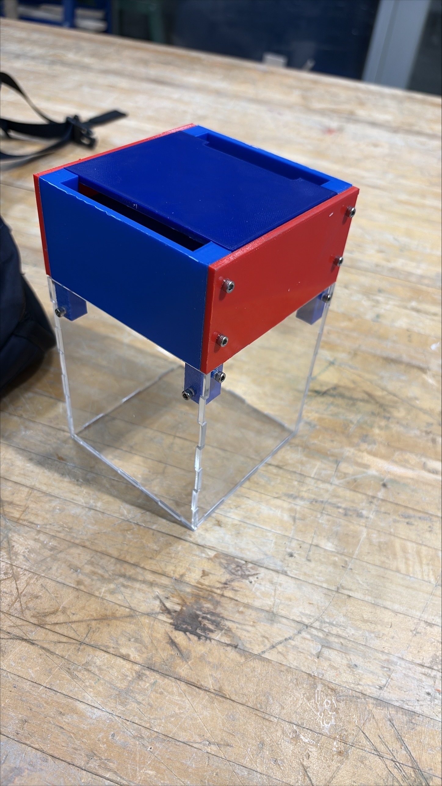 The gimbal and camera enclosure assembled: a red and blue printed frame bolted on top of a clear acrylic housing, standing on a workbench.